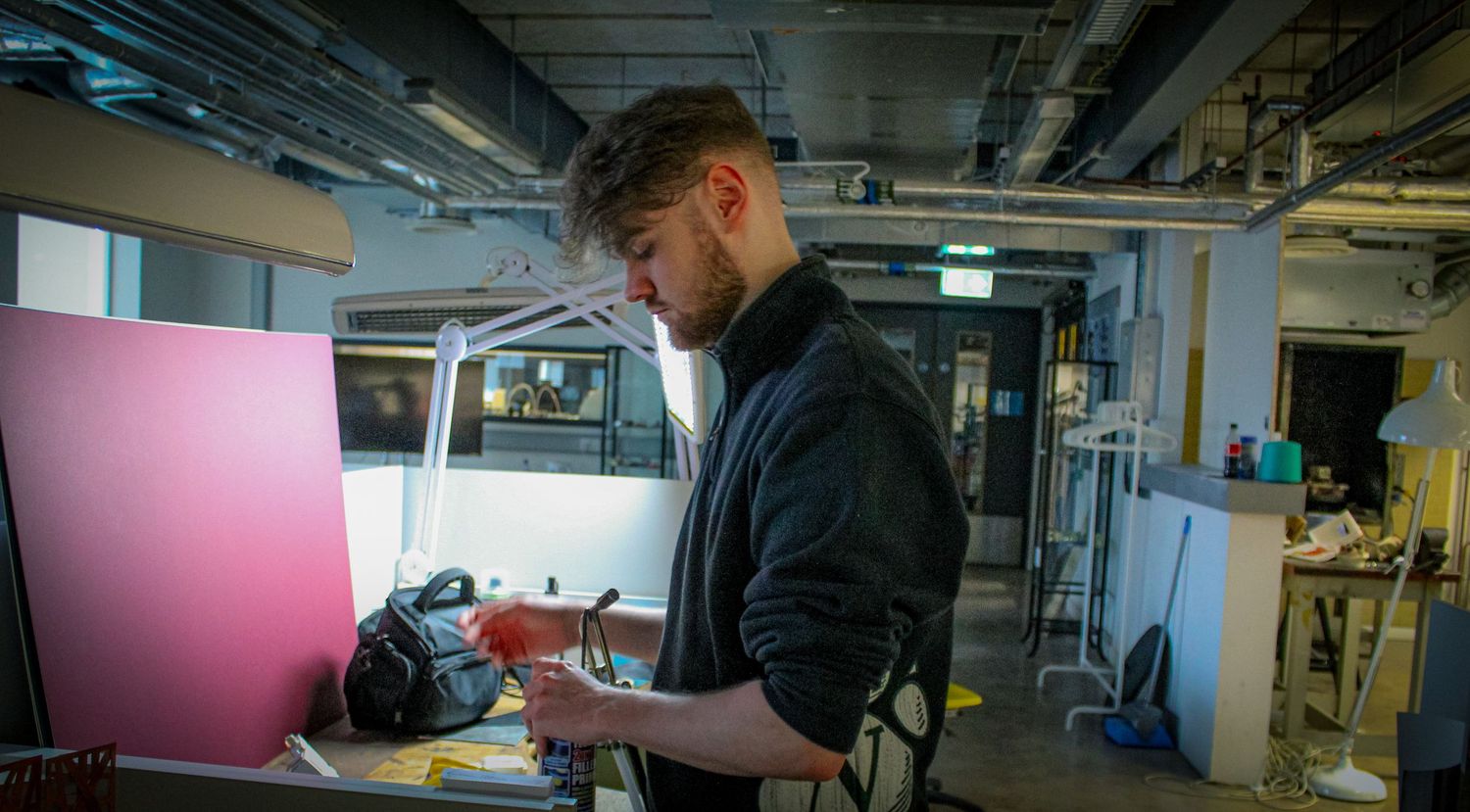 A young man in a black jacket works at a cluttered workbench in a modern industrial studio space.