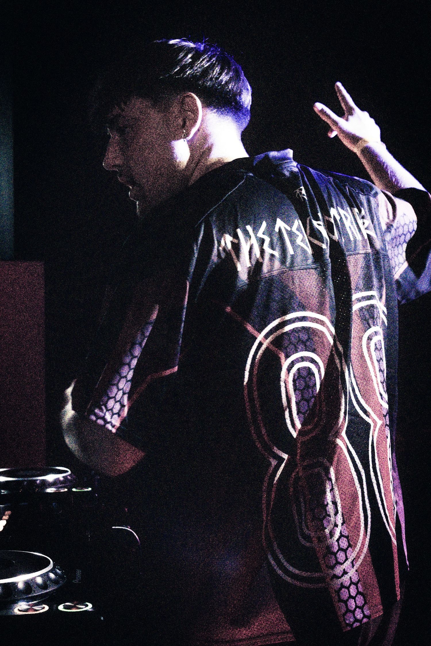 DJ performing at a dark nightclub venue wearing a decorative jersey with circular patterns under blue stage lighting.