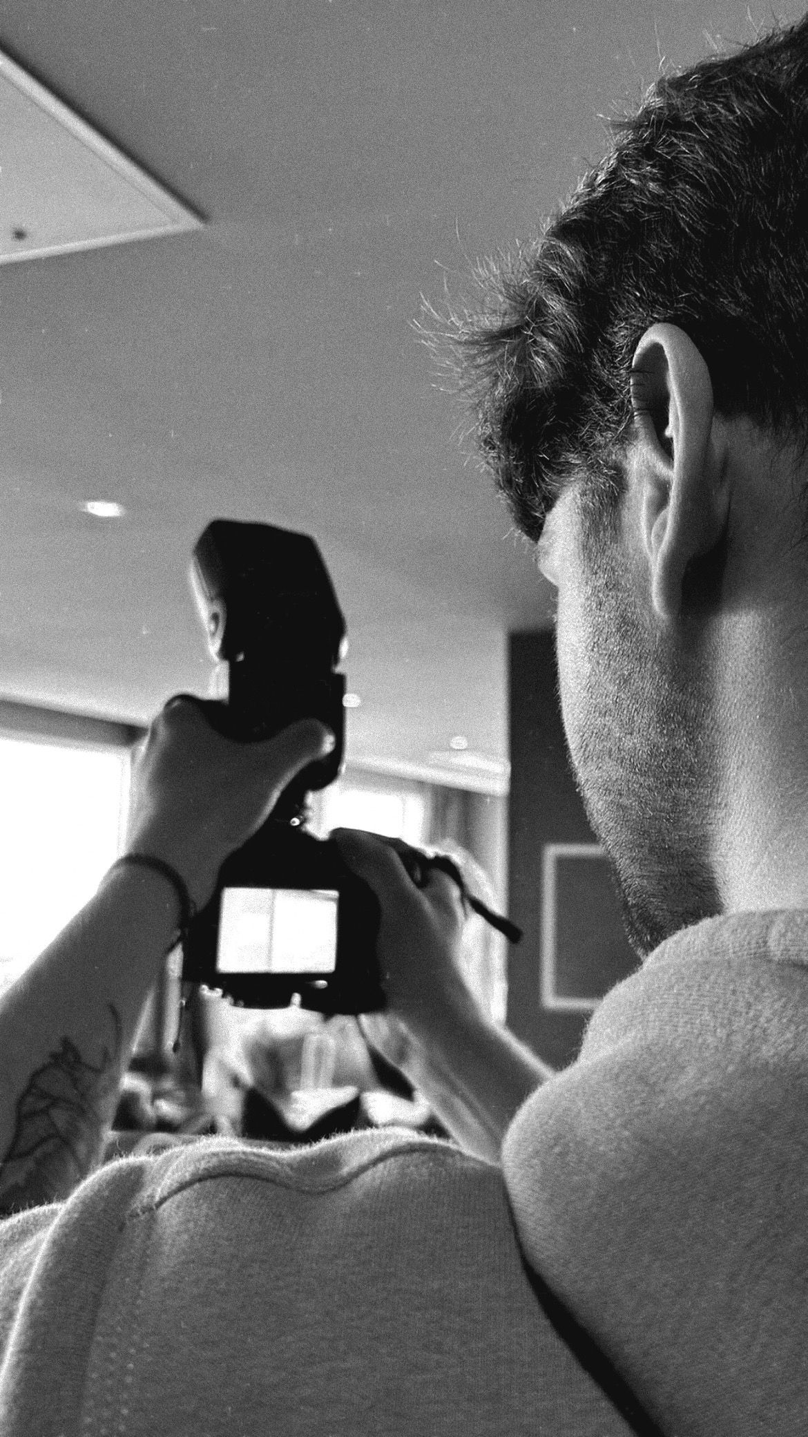 Black and white photo of a man holding a camera with flash, viewed from behind, with a tattoo visible on his wrist.