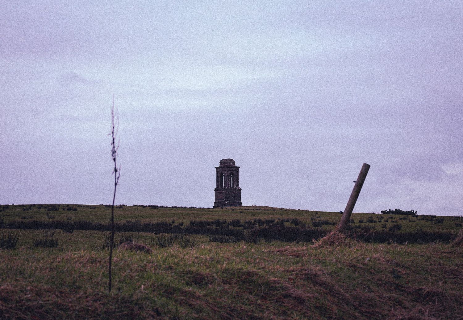 A stone tower monument stands on a grassy hilltop under an overcast sky with a wooden fence post in the foreground.
