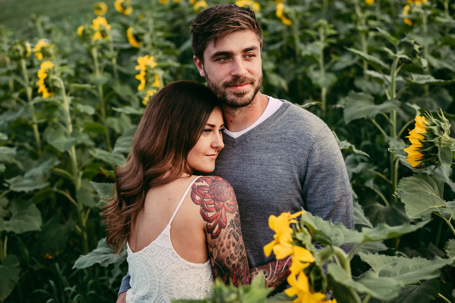 Romantic couple embracing in a vibrant sunflower field, woman with sleeve tattoos resting on mans chest.