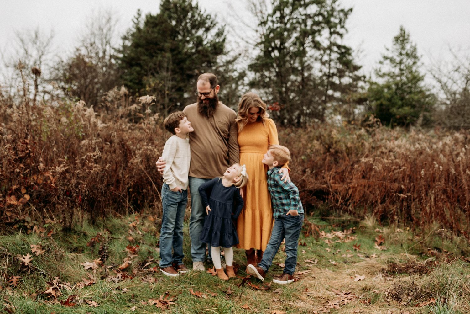 A loving family of five huddles together on a woodland path surrounded by golden autumn brush and trees.