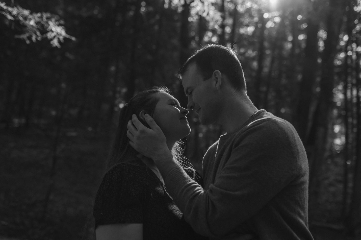 Black and white romantic photo of couple nearly kissing in sunlit forest, faces close together.