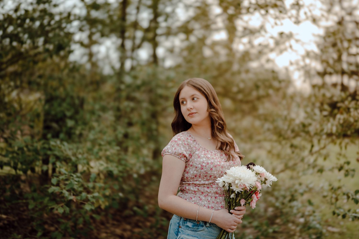 Young woman holding white flowers standing in a lush green forest path during golden hour light.