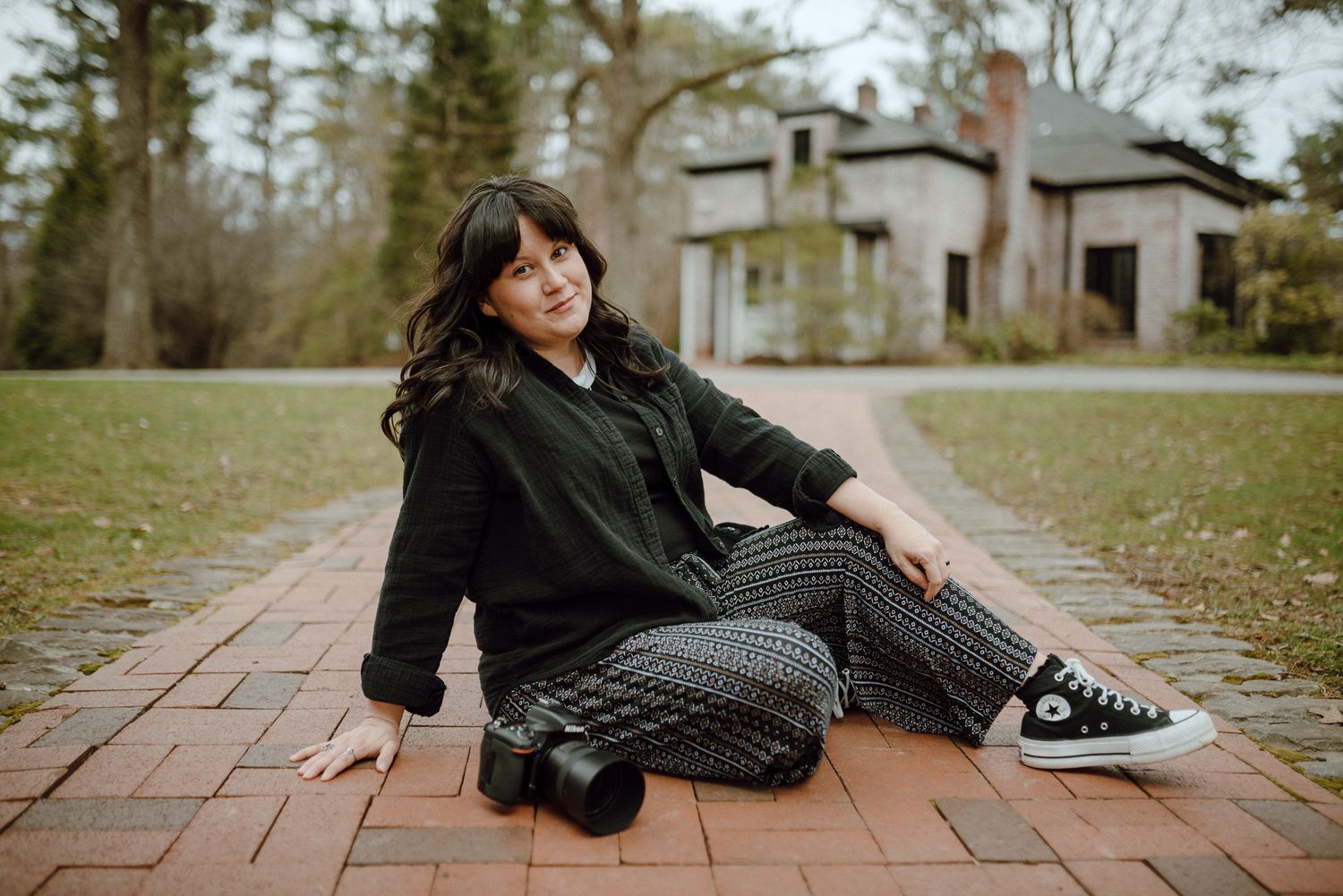 Female photographer sits on brick pathway holding camera, wearing black jacket and patterned pants near historic building.