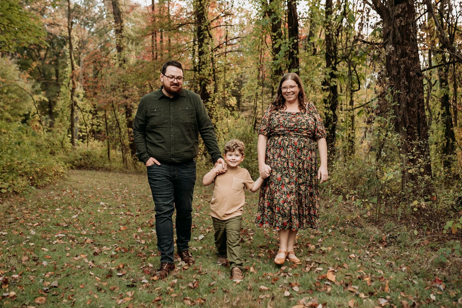 A smiling family of three walks through a colorful autumn forest, holding hands among fallen leaves.