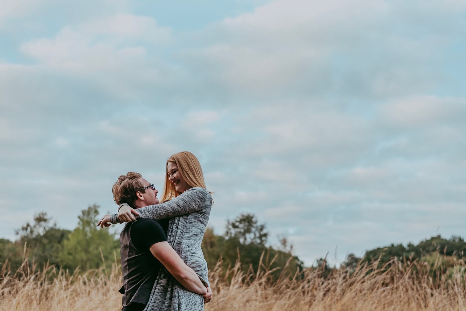 Couple embracing in a golden field under a cloudy sky during engagement shoot.