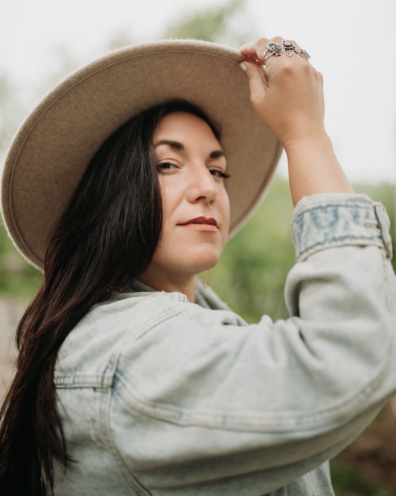 Brunette woman in denim jacket holds wide-brim hat outdoors, wearing silver ring in soft natural light.