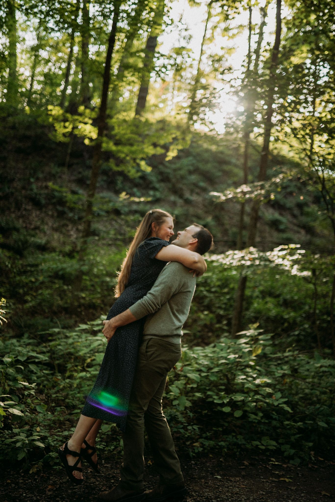 A couple shares a joyful embrace as the man lifts his partner in a sun-dappled green forest setting. Engagement session.
