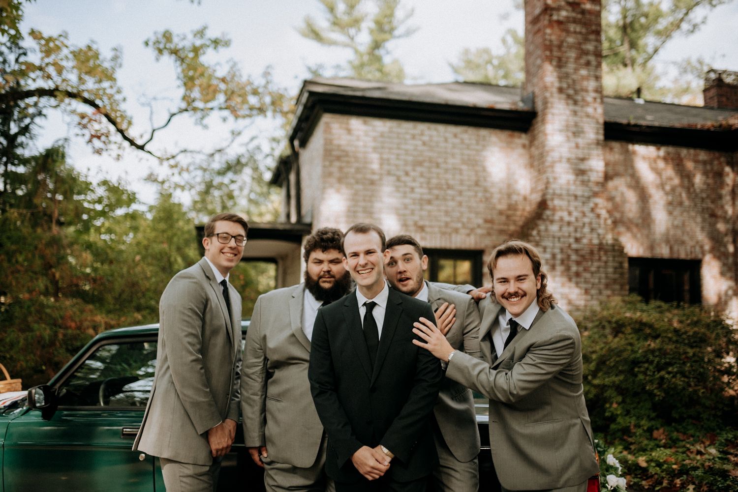 Five groomsmen in gray suits posing and smiling beside a vintage green car outside brick house.