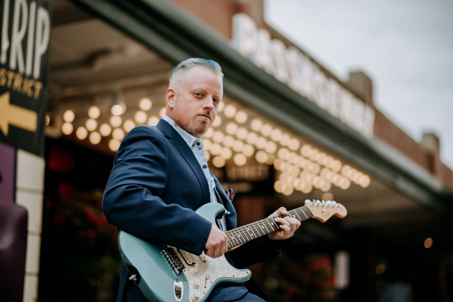Man in blue suit playing electric guitar outside lit marquee sign on urban street. Musician creative portrait.
