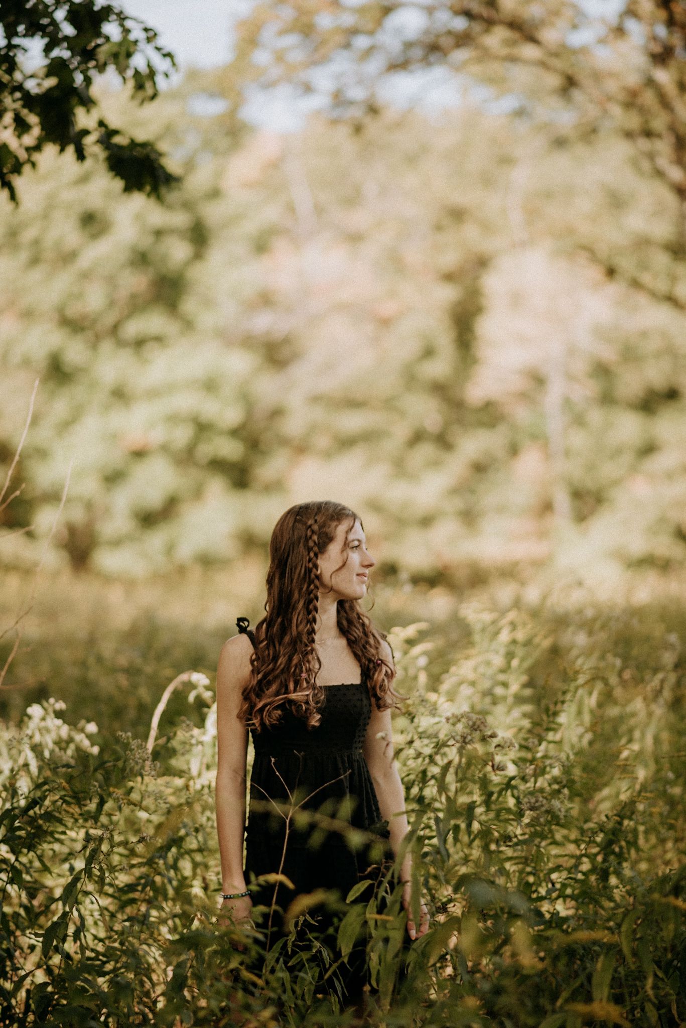 Young woman with long curly hair in a black dress standing in a sunlit wildflower meadow. Senior Portrait Photography.