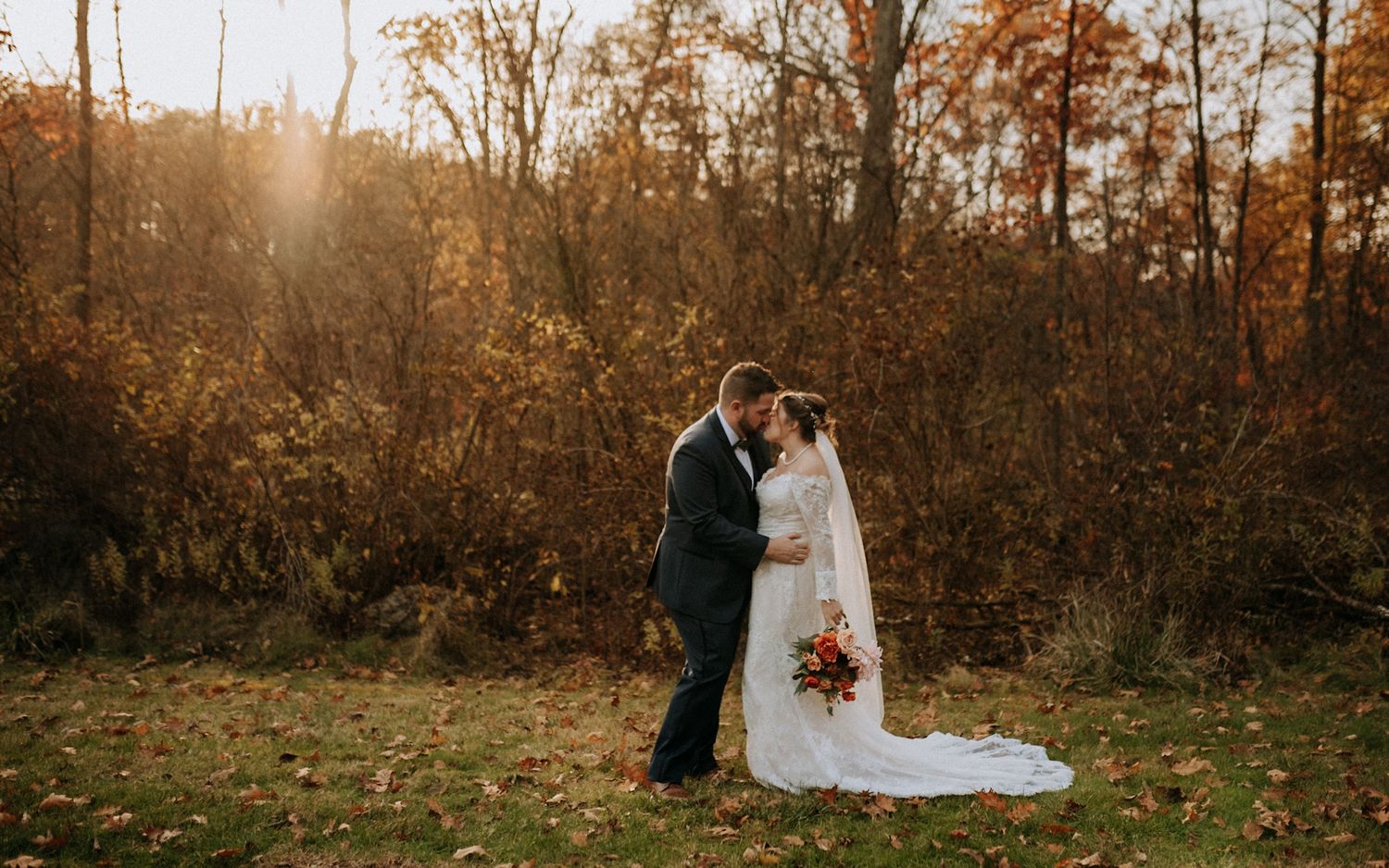 Bride and groom kissing in golden autumn field, she holds red bouquet, surrounded by fall foliage.