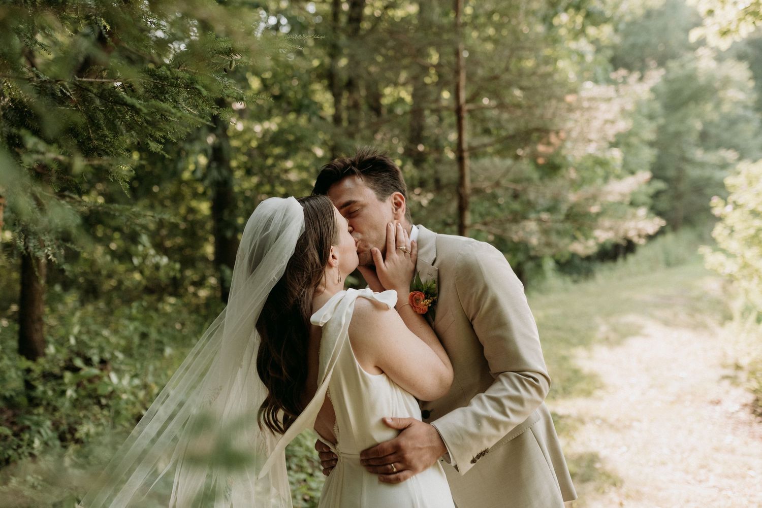 Bride and groom sharing a romantic kiss in a sun-dappled forest on their wedding day.