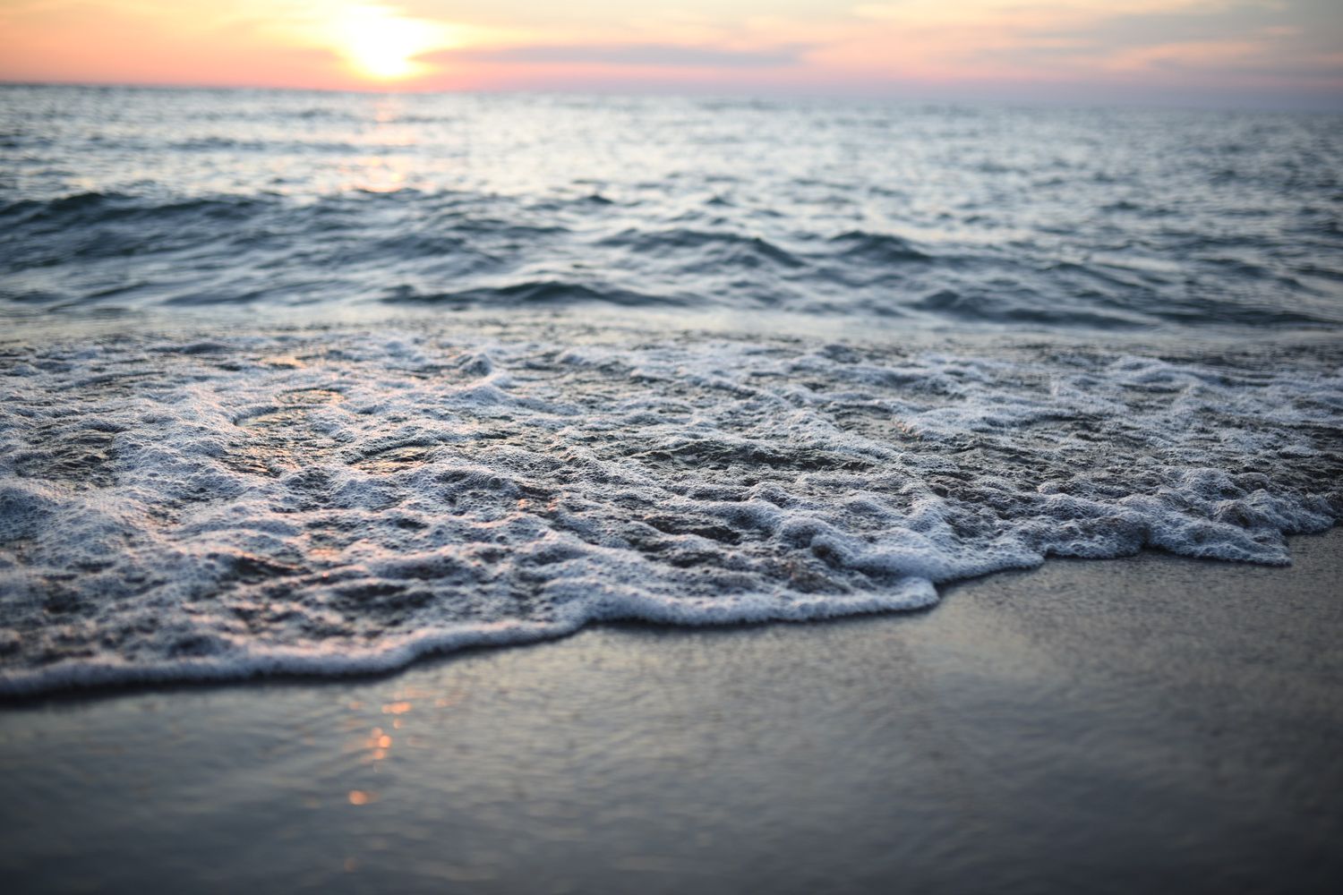 Soft ocean waves rolling onto a sandy shore at sunset with warm pink and orange sky reflections.