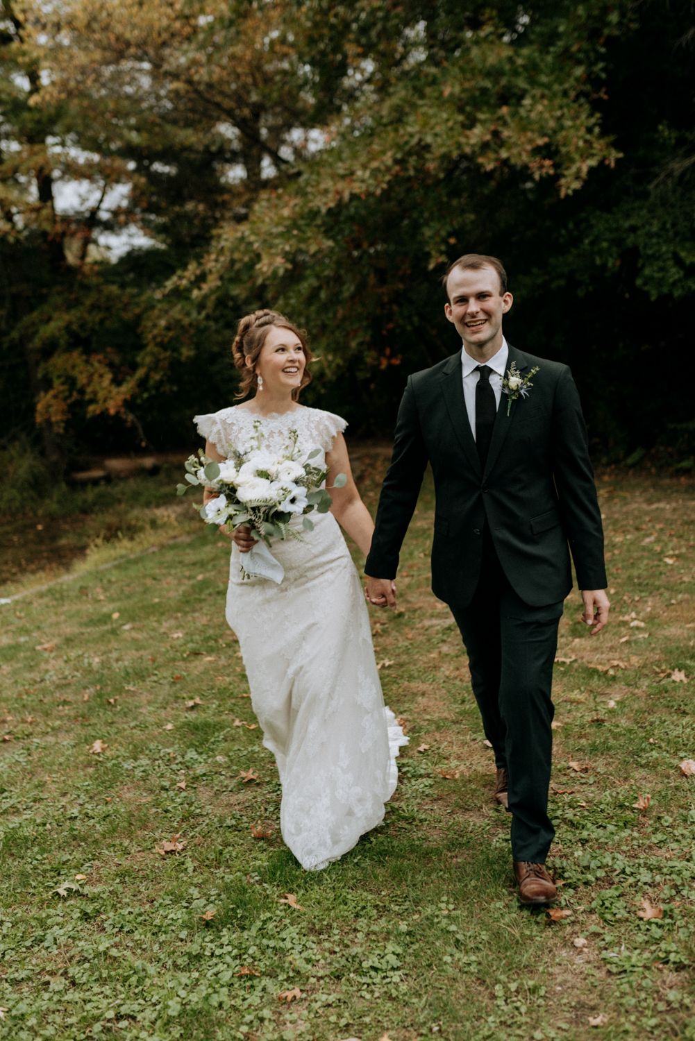 Smiling bride and groom walk hand-in-hand through green lawn surrounded by autumn trees, bride holding white bouquet.