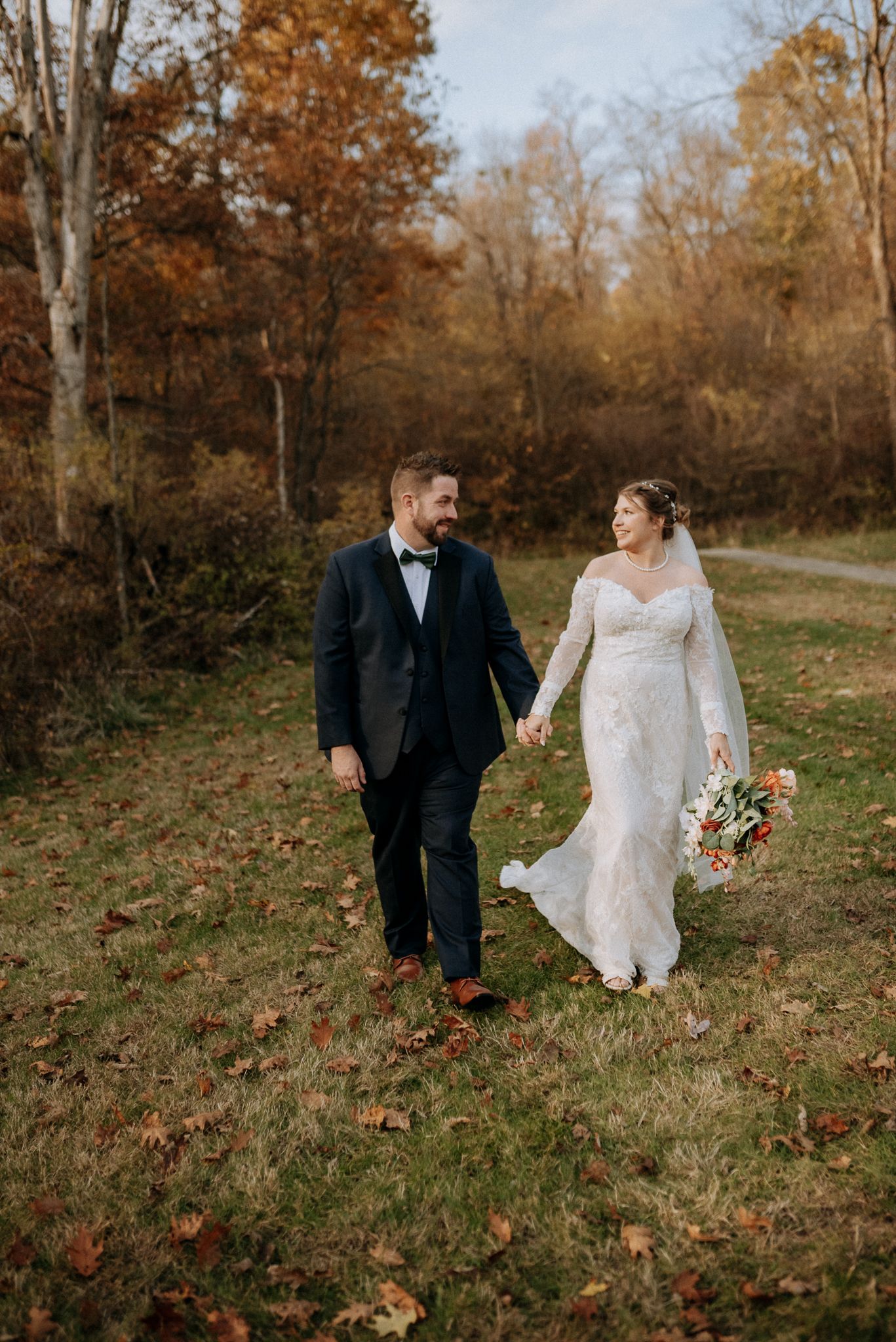 Bride and groom walking hand in hand through a colorful autumn forest in wedding attire.