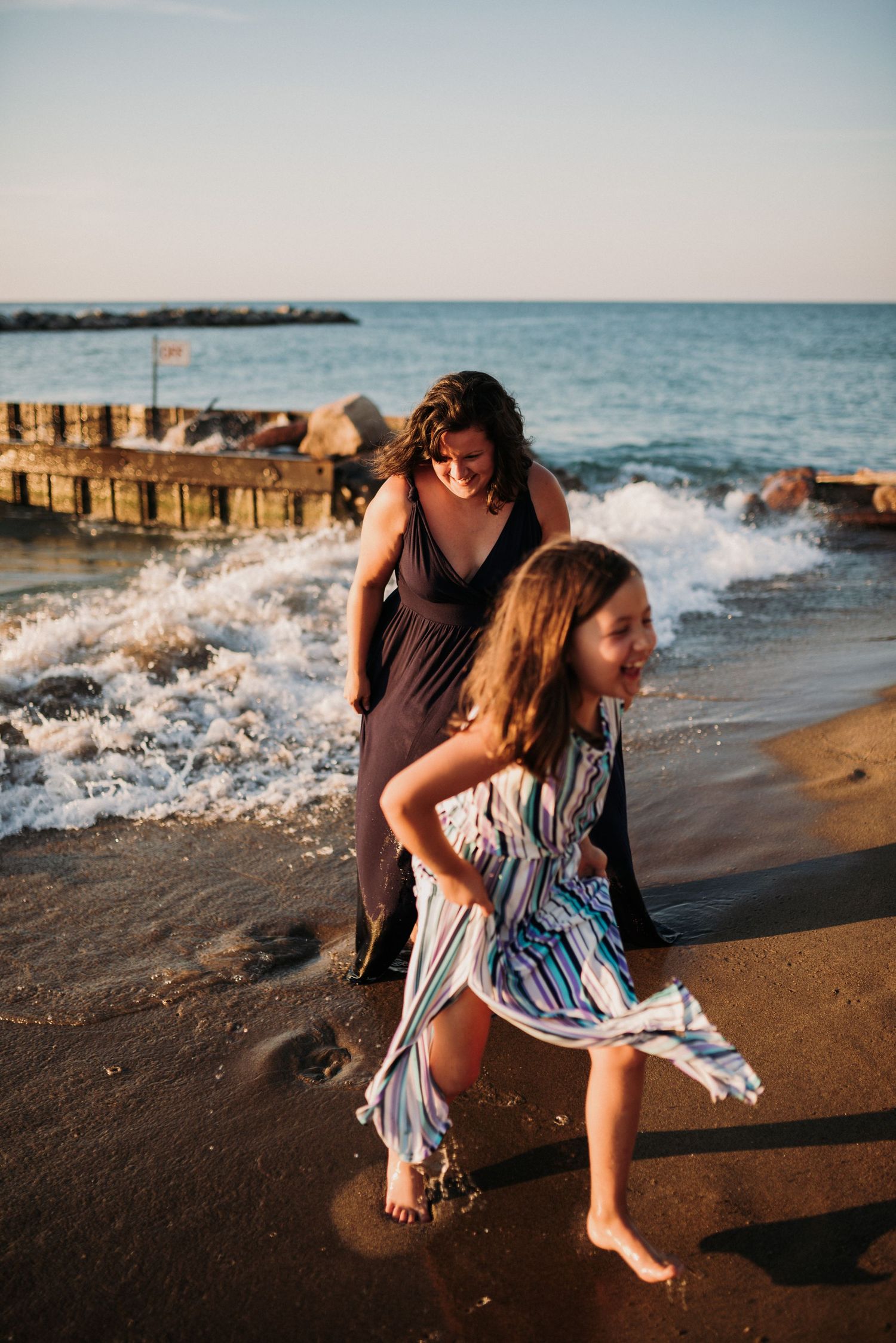 Mother and daughter laughing and playing together on a sandy beach with ocean waves in the background.