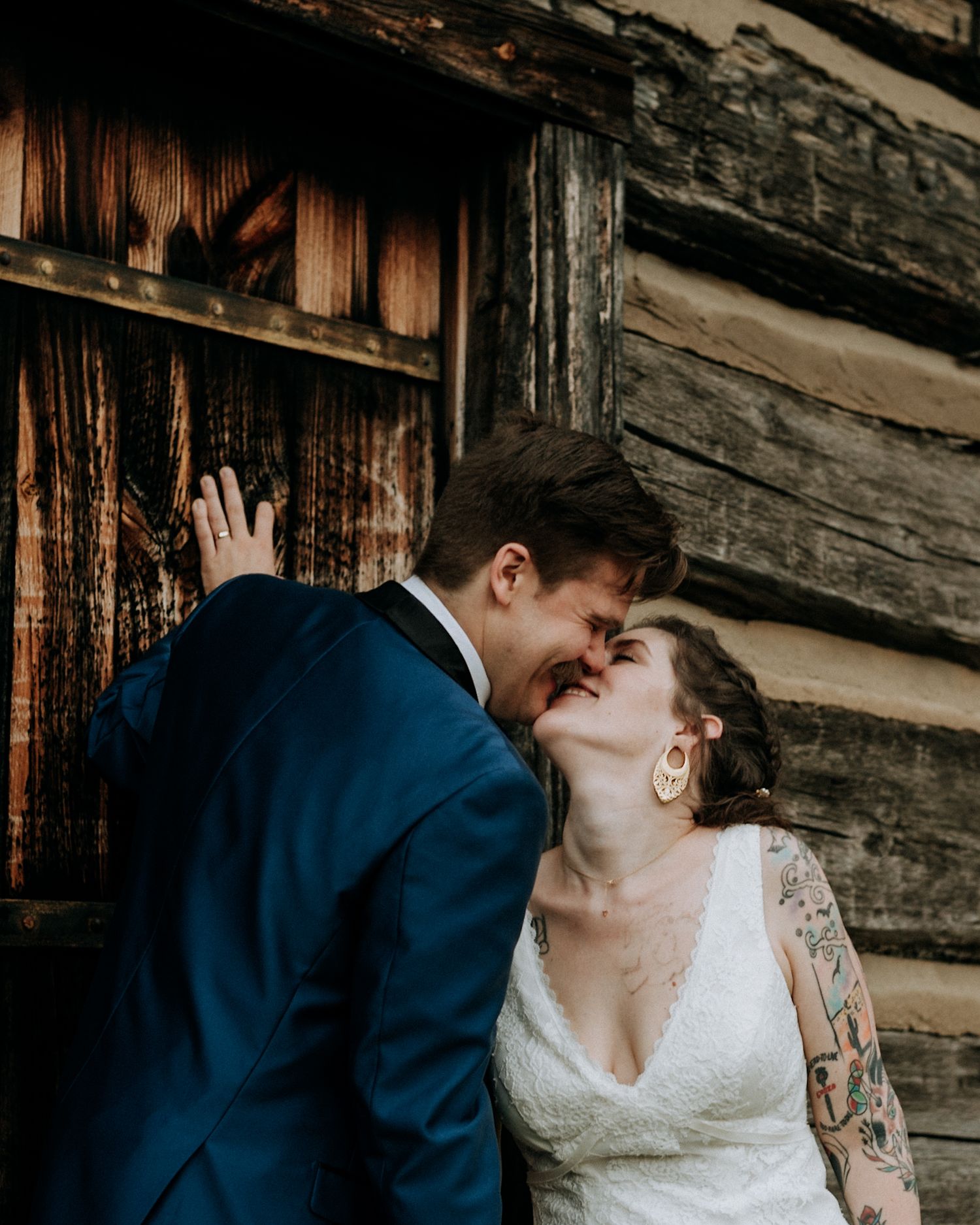 Bride and groom share an intimate laughing moment together leaning against a rustic log cabin wall during their wedding.