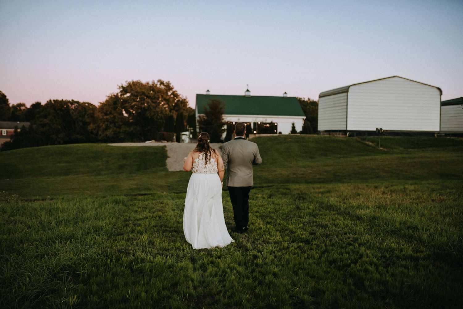 Bride and groom walking hand in hand across green field toward white barn at sunset.