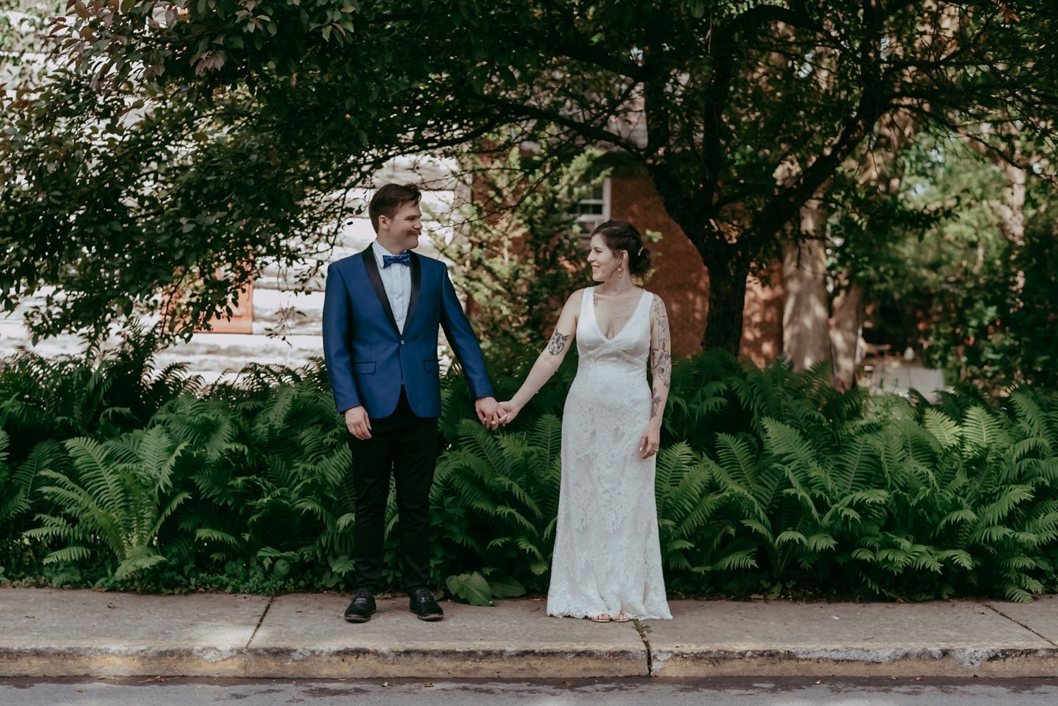 Bride and groom hold hands smiling outdoors surrounded by lush green ferns on their wedding day.