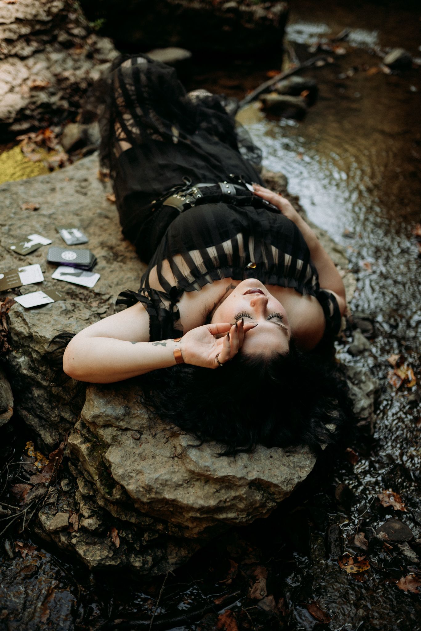 A woman in a black dress lies dramatically on rocks near a stream with tarot cards scattered nearby.