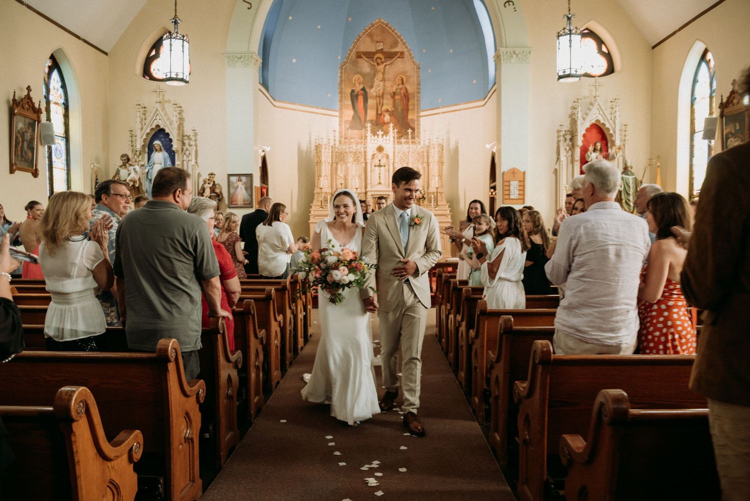 Newlyweds walk down church aisle after ceremony, bride holds bouquet, guests celebrate in ornate Catholic church interior.