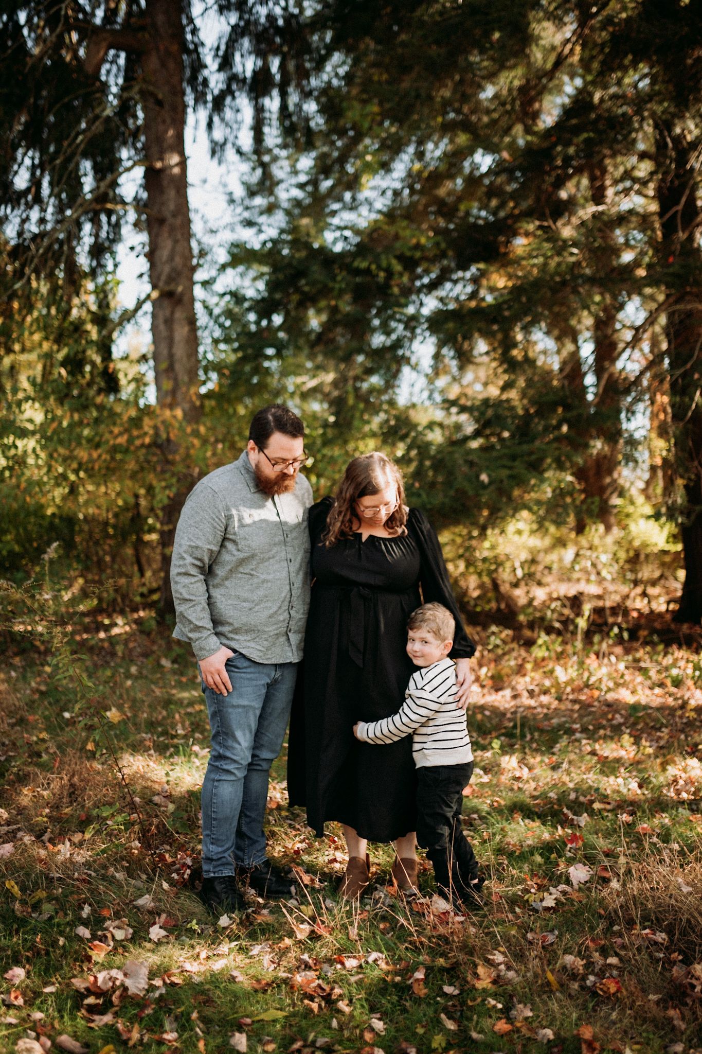 A family of three enjoys a sunny autumn day in the woods, with parents smiling as toddler reaches up to mom. Fall mini sessions in Butler.