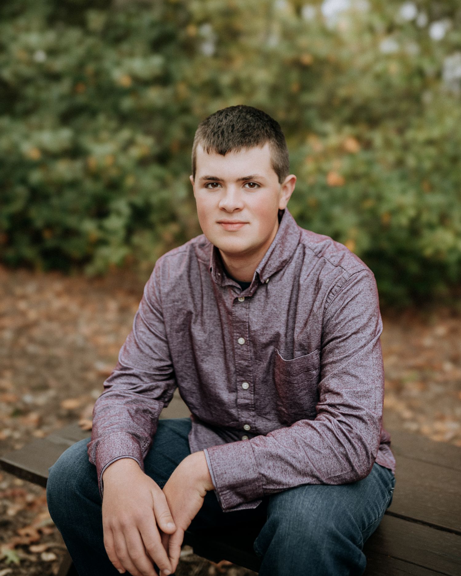Young man in purple button-up shirt sitting outdoors for fall senior portrait photography session.