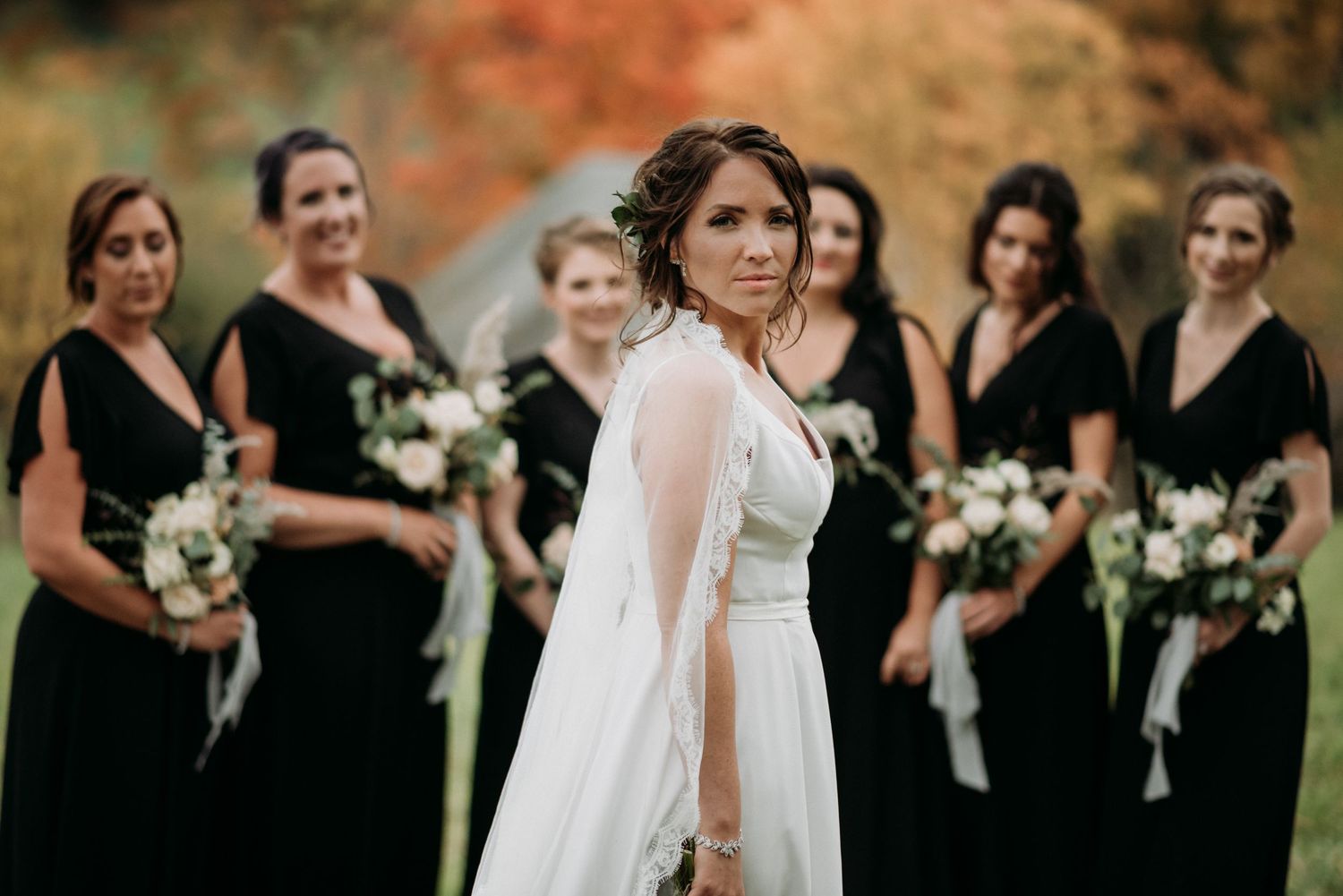 Bride in flowing white gown poses with bridesmaids dressed in black holding white floral bouquets.