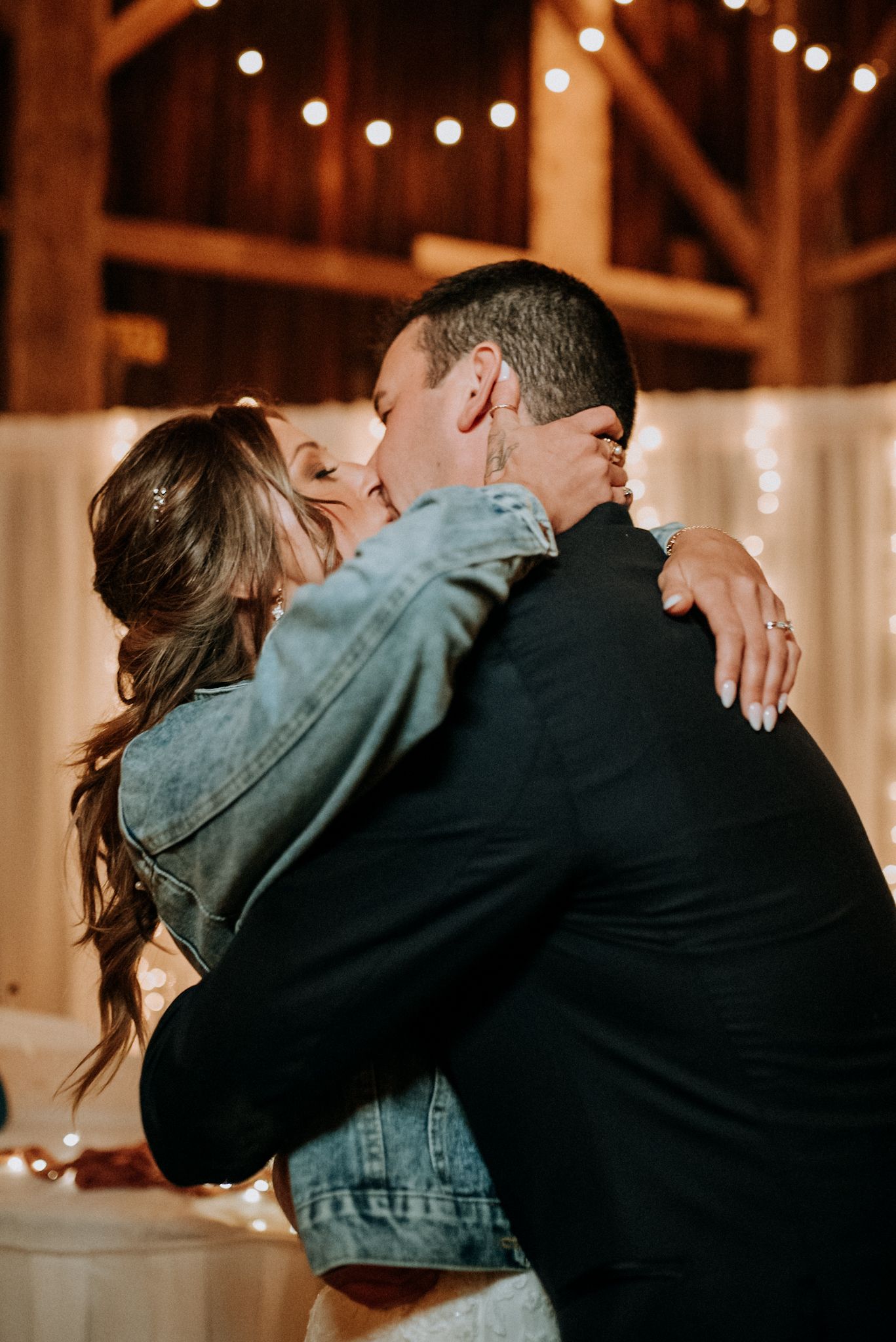 A bride in a denim jacket and groom share a passionate kiss under warm string lights in a rustic barn. Butler County, Pennsylvania wedding.