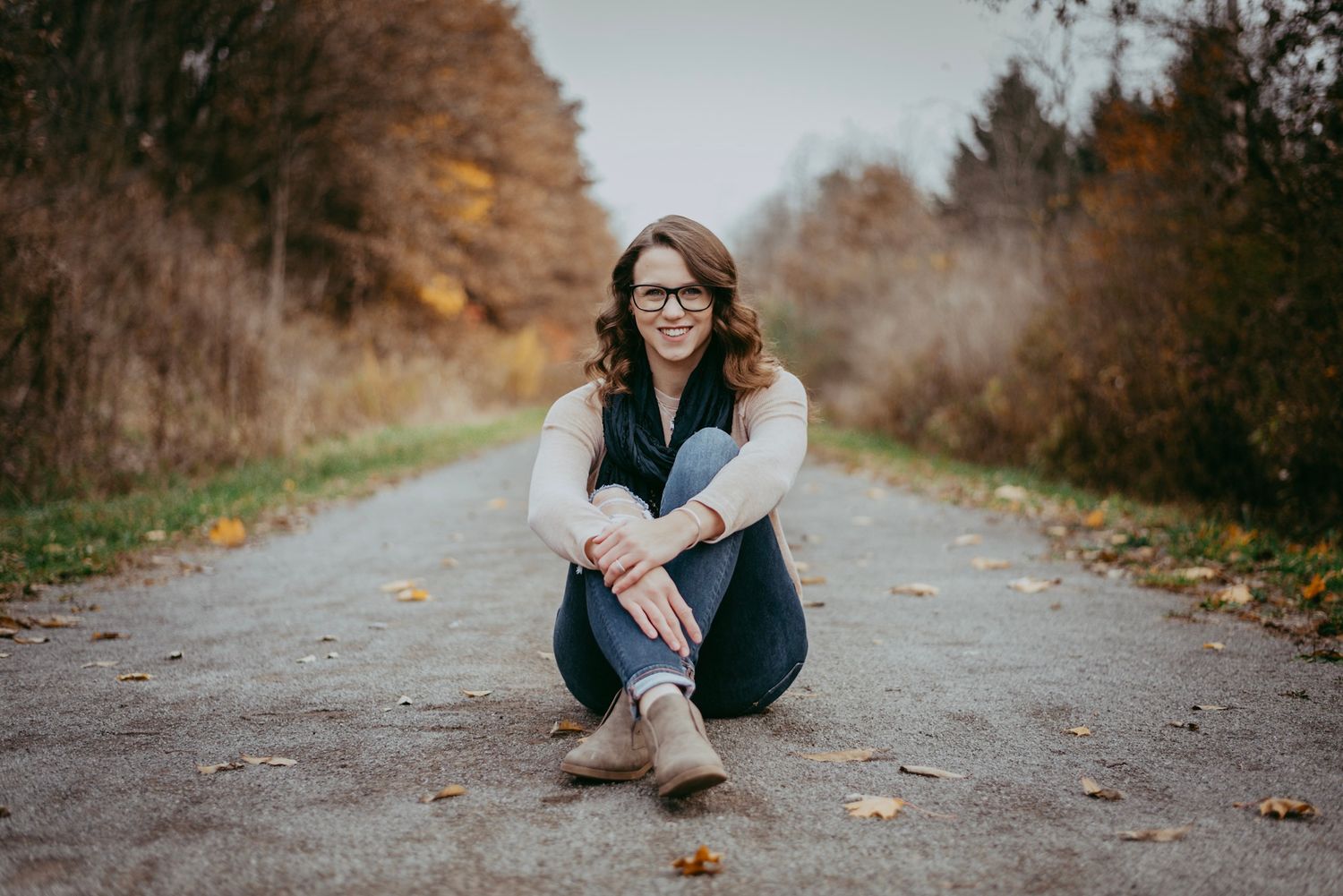 A smiling young woman with glasses sits on a tree-lined path surrounded by autumn foliage in a casual outdoor senior portrait.