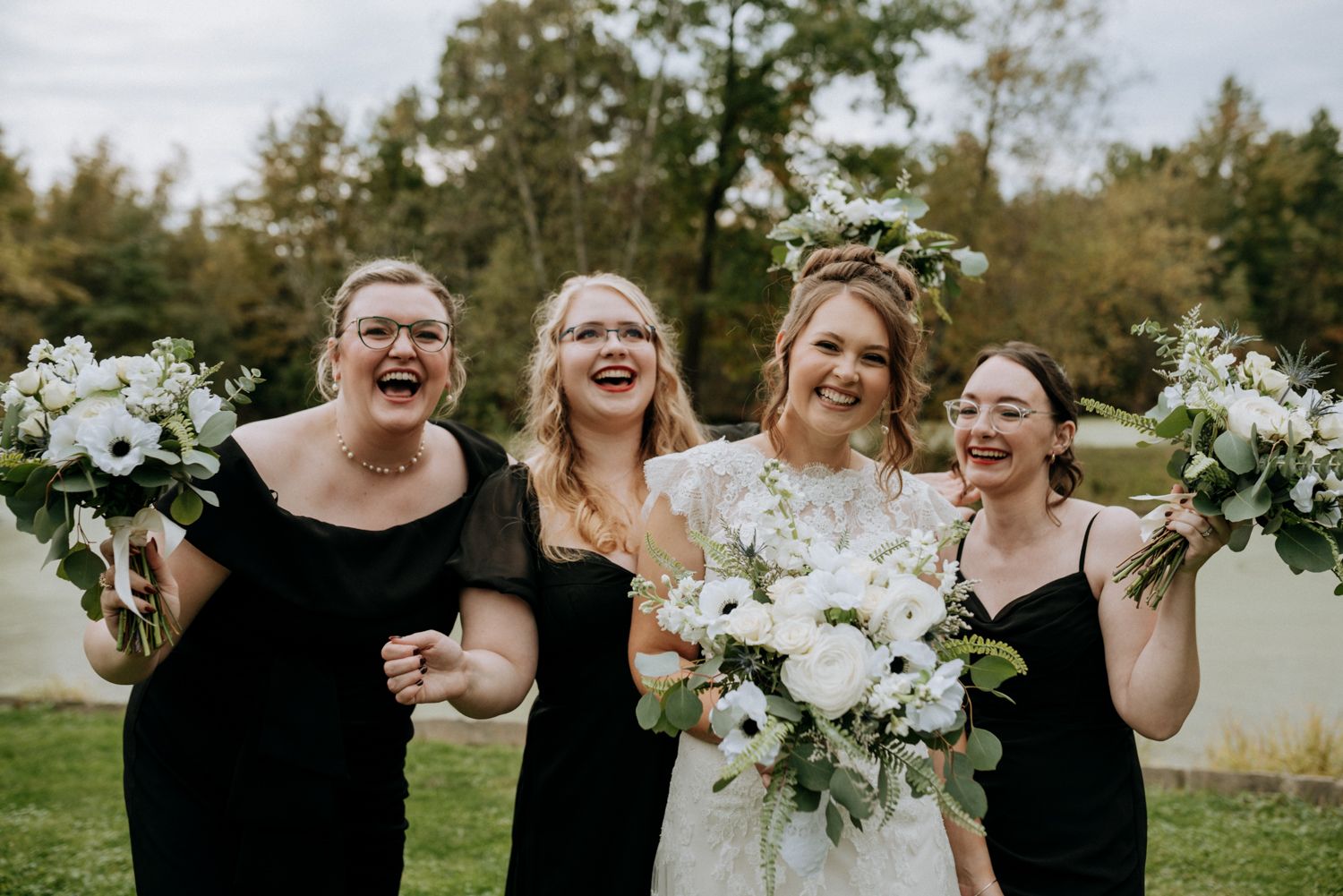 Bride in white lace gown with floral crown laughs with three bridesmaids in black dresses holding white bouquets outdoors.