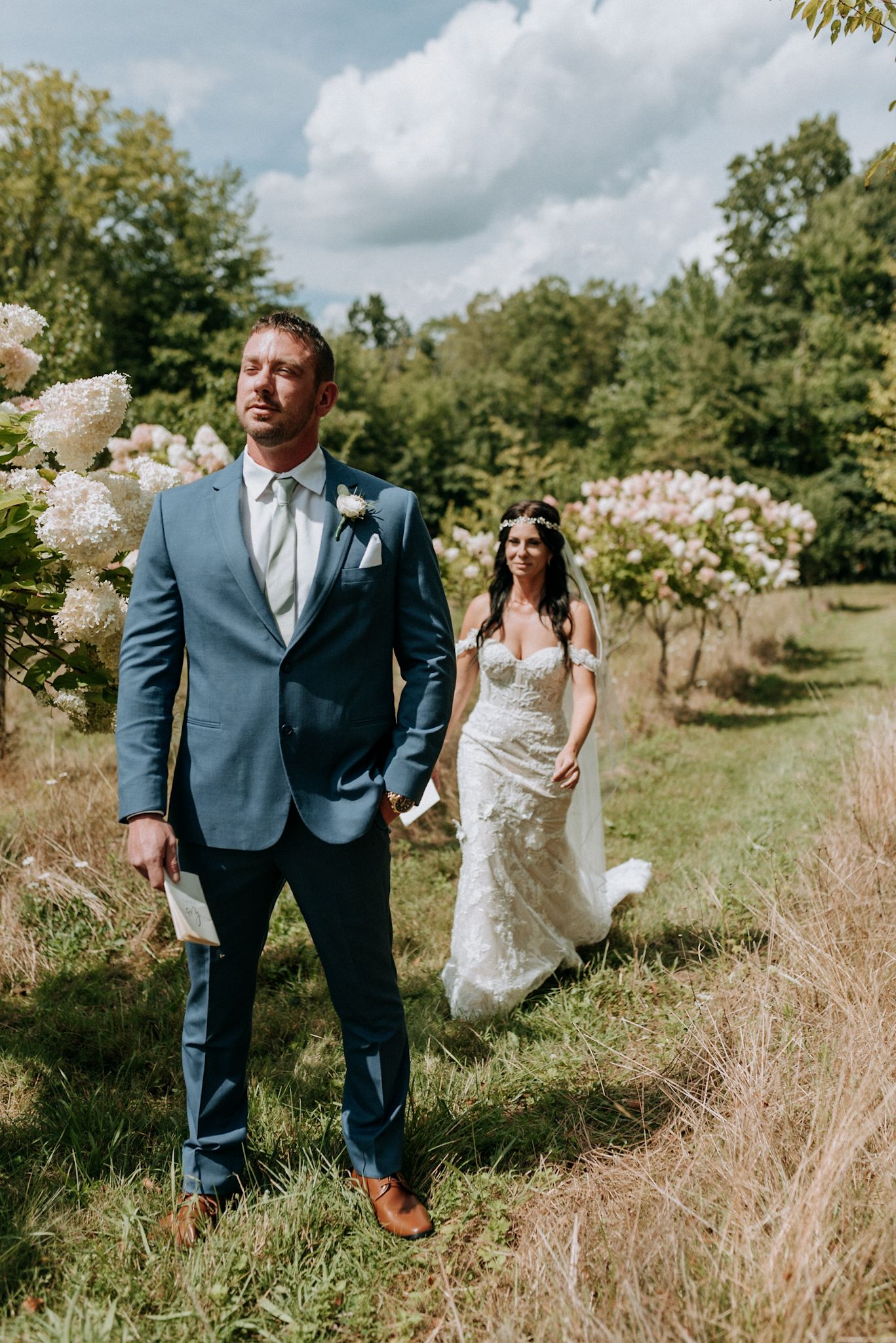 Bride in lace gown approaching groom in blue suit during outdoor first look ceremony.