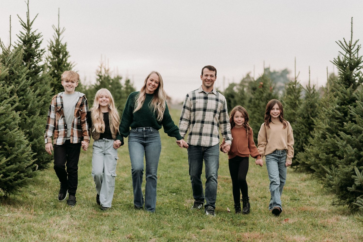 Family of six walking hand in hand through Christmas tree farm on overcast day.