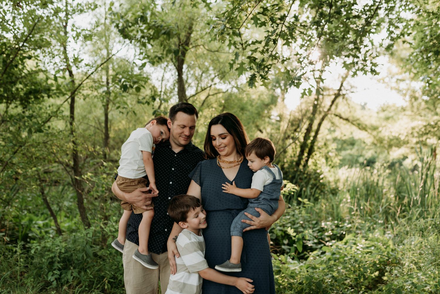 A family of five poses outdoors among lush green trees, with parents holding young children in a warm, natural setting.