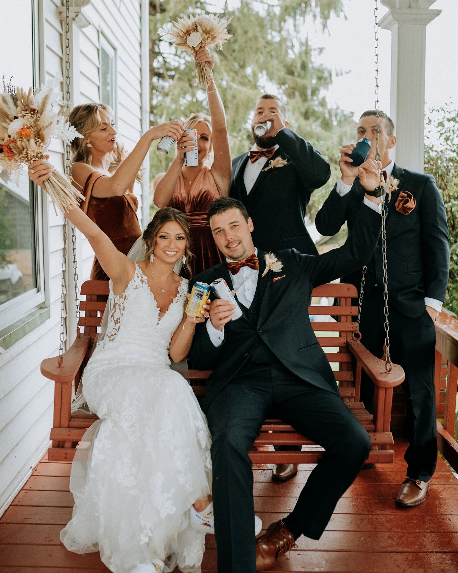 Excited wedding party celebrates on a porch swing, bride and groom toasting with bridesmaids and groomsmen in festive attire.