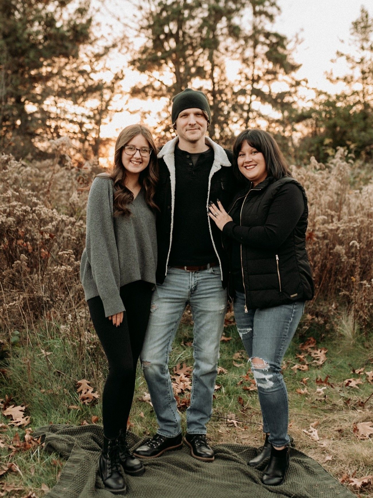 Three people standing together on a blanket in a fall forest, smiling during a golden sunset.