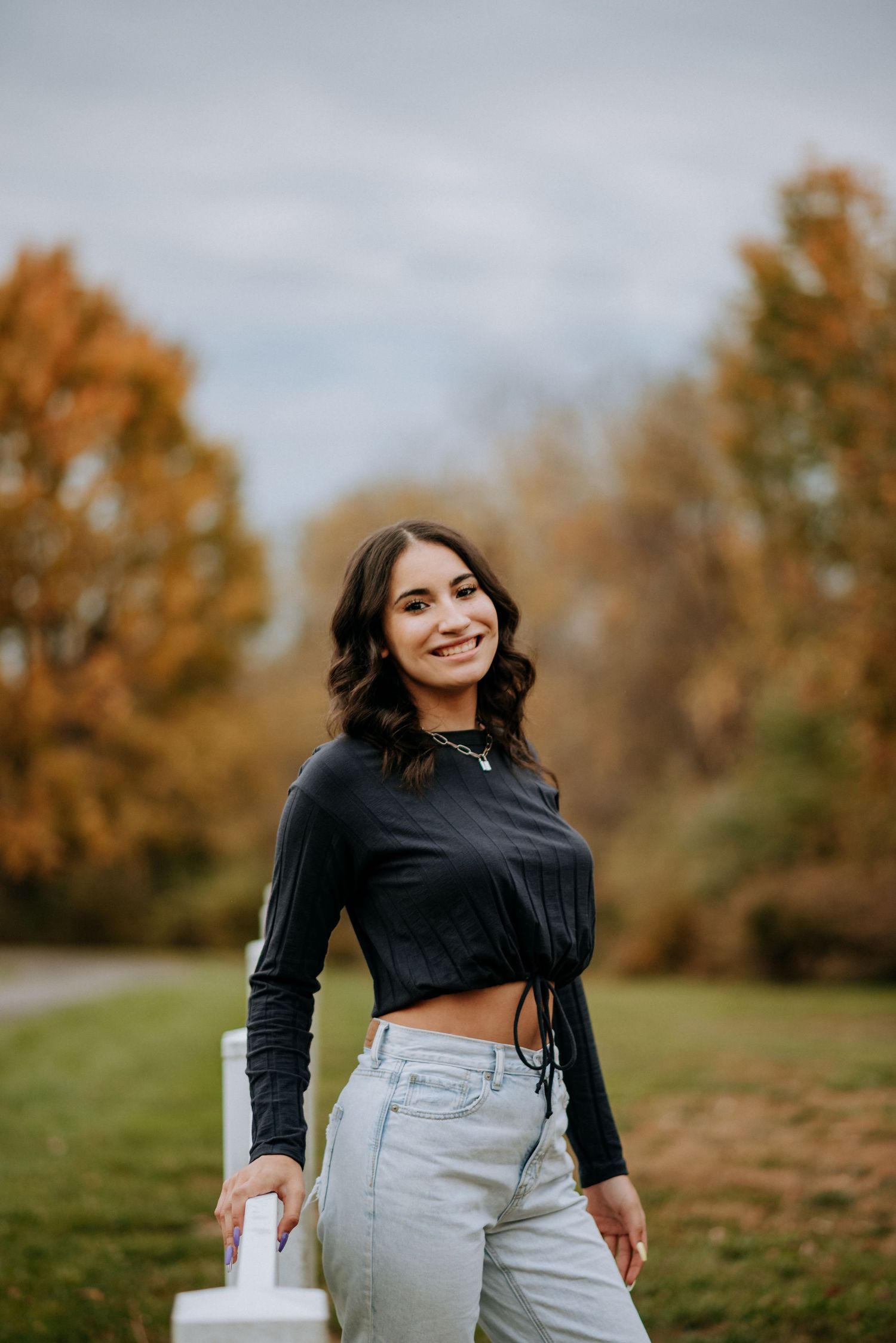 Young woman in black crop top and jeans smiling outdoors during autumn fall season senior portrait photography.