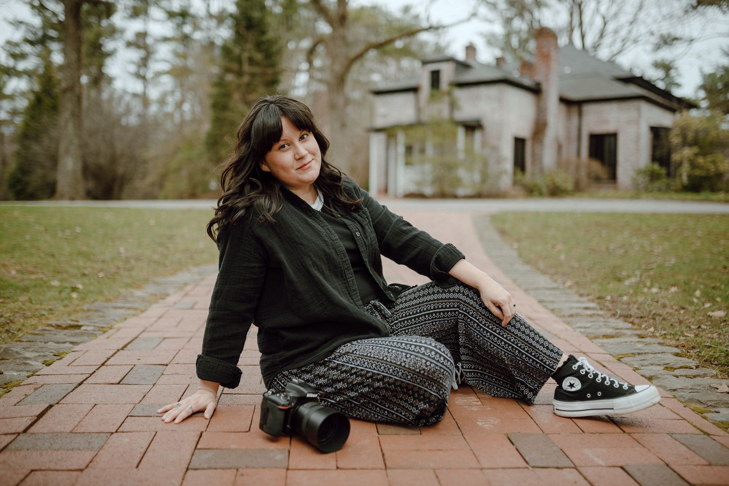 Trista Renee Photography sitting on brick path holding camera, wearing black jacket and plaid pants with Converse sneakers outdoors.