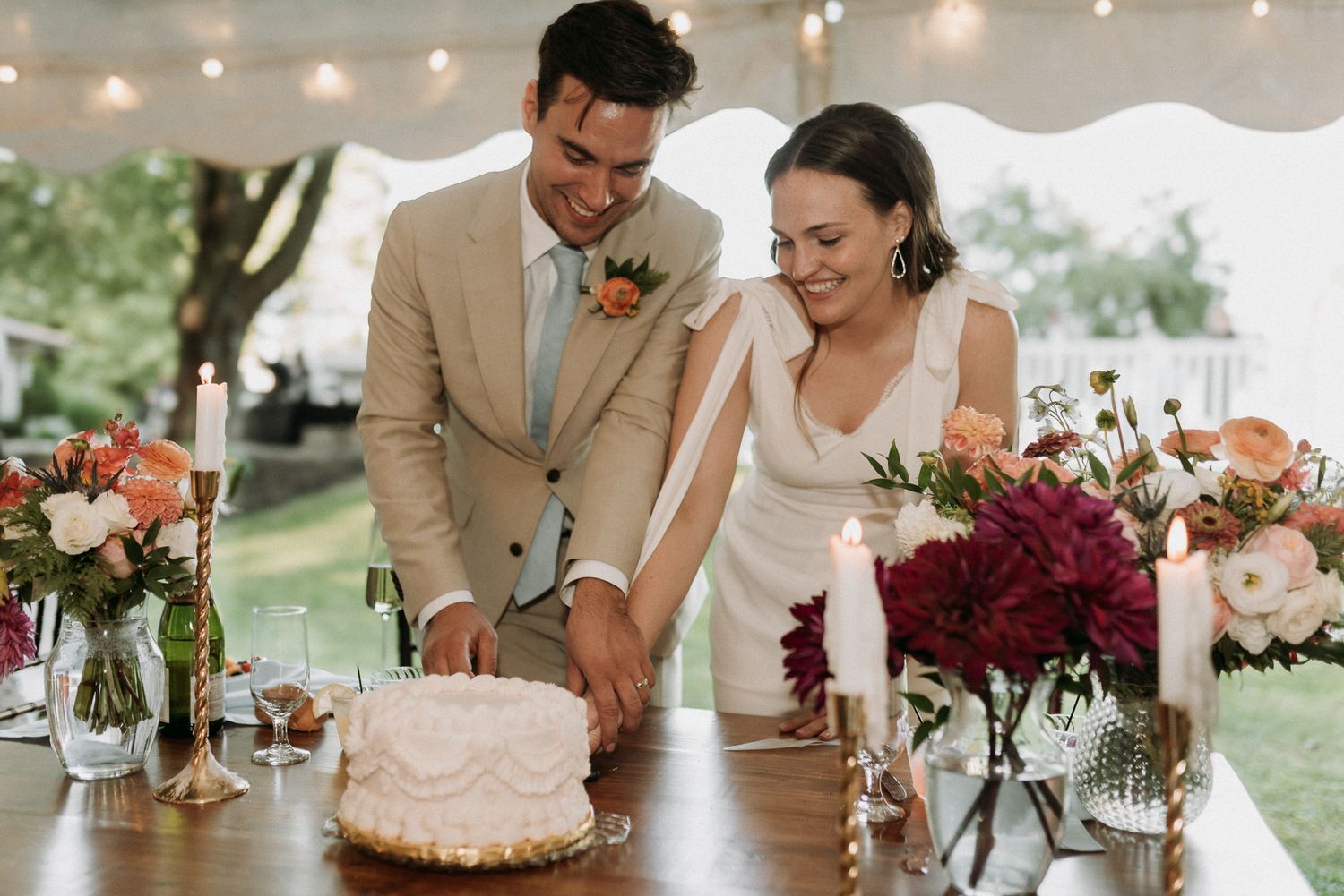 A smiling couple cuts their wedding cake under a tent surrounded by colorful floral arrangements.