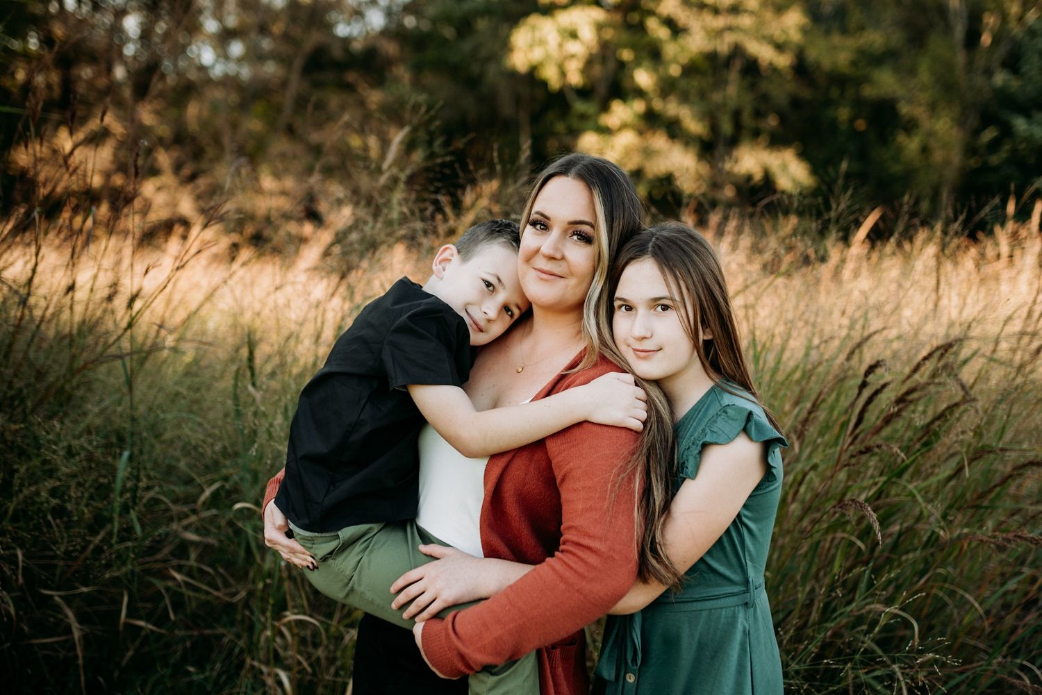 A mother tenderly embraces her two children in a sunlit field of tall golden grass during autumn.