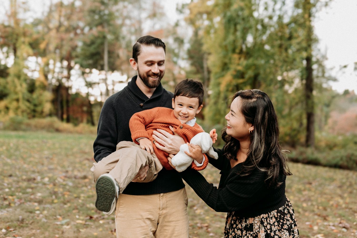 A joyful family of three poses outdoors in autumn, with dad holding toddler while mom smiles in a fall park setting.