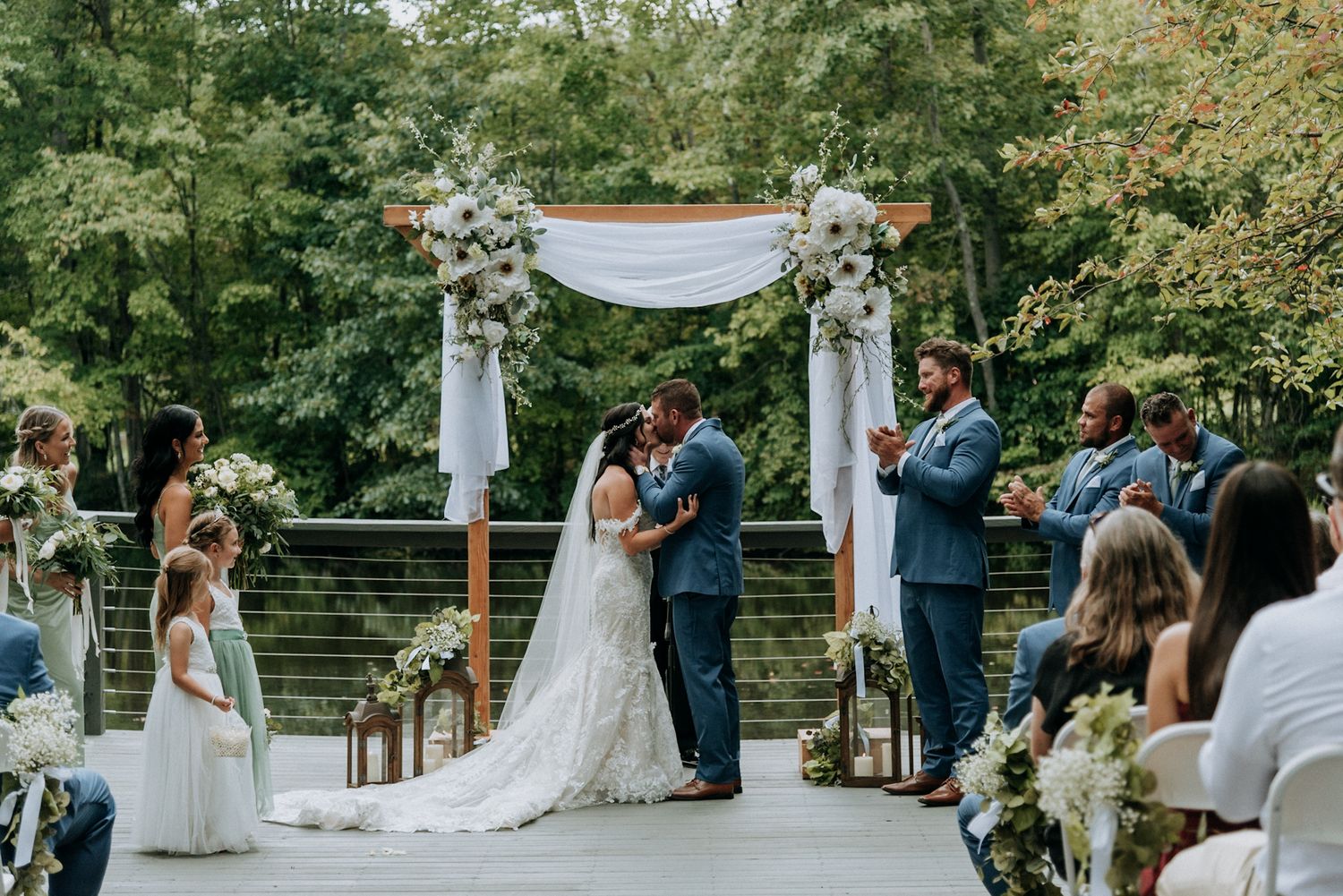 Bride and groom share a first kiss at an elegant outdoor wedding ceremony beneath a floral arch surrounded by guests.