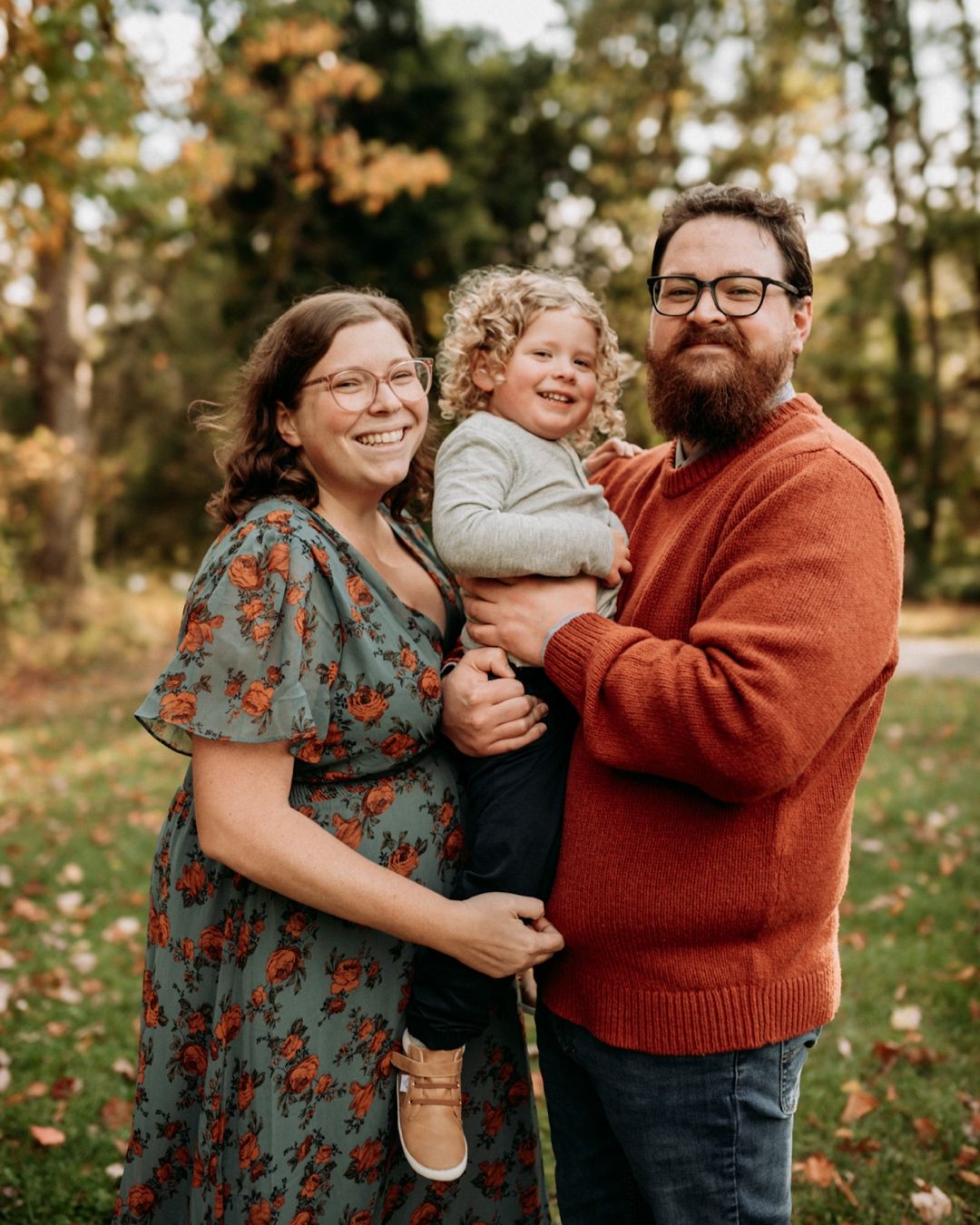A smiling family of three poses outdoors in fall foliage, parents holding a young curly-haired toddler.