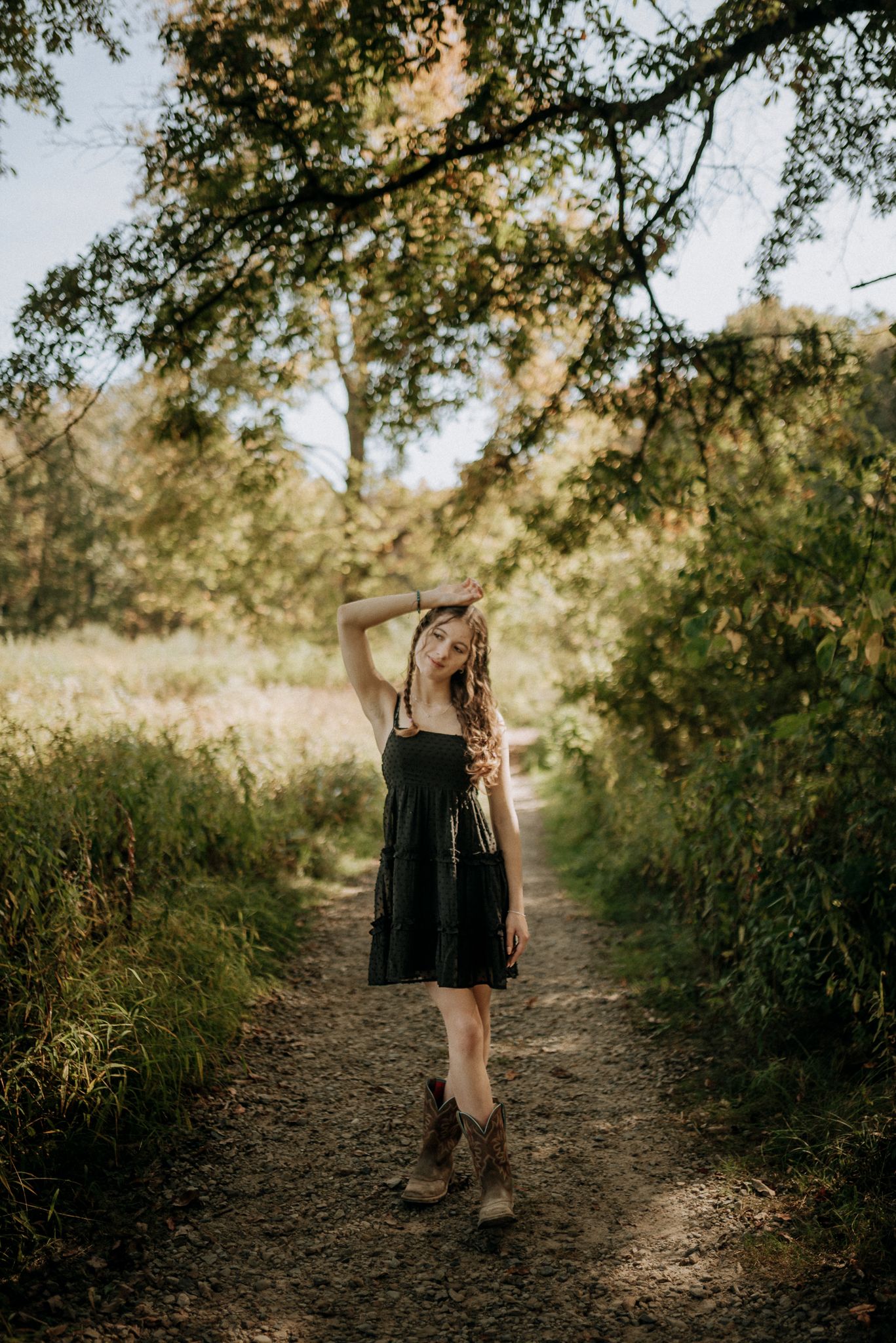 A young woman in a black dress and cowboy boots poses on a dirt path surrounded by lush greenery. Senior Portrait Photography.