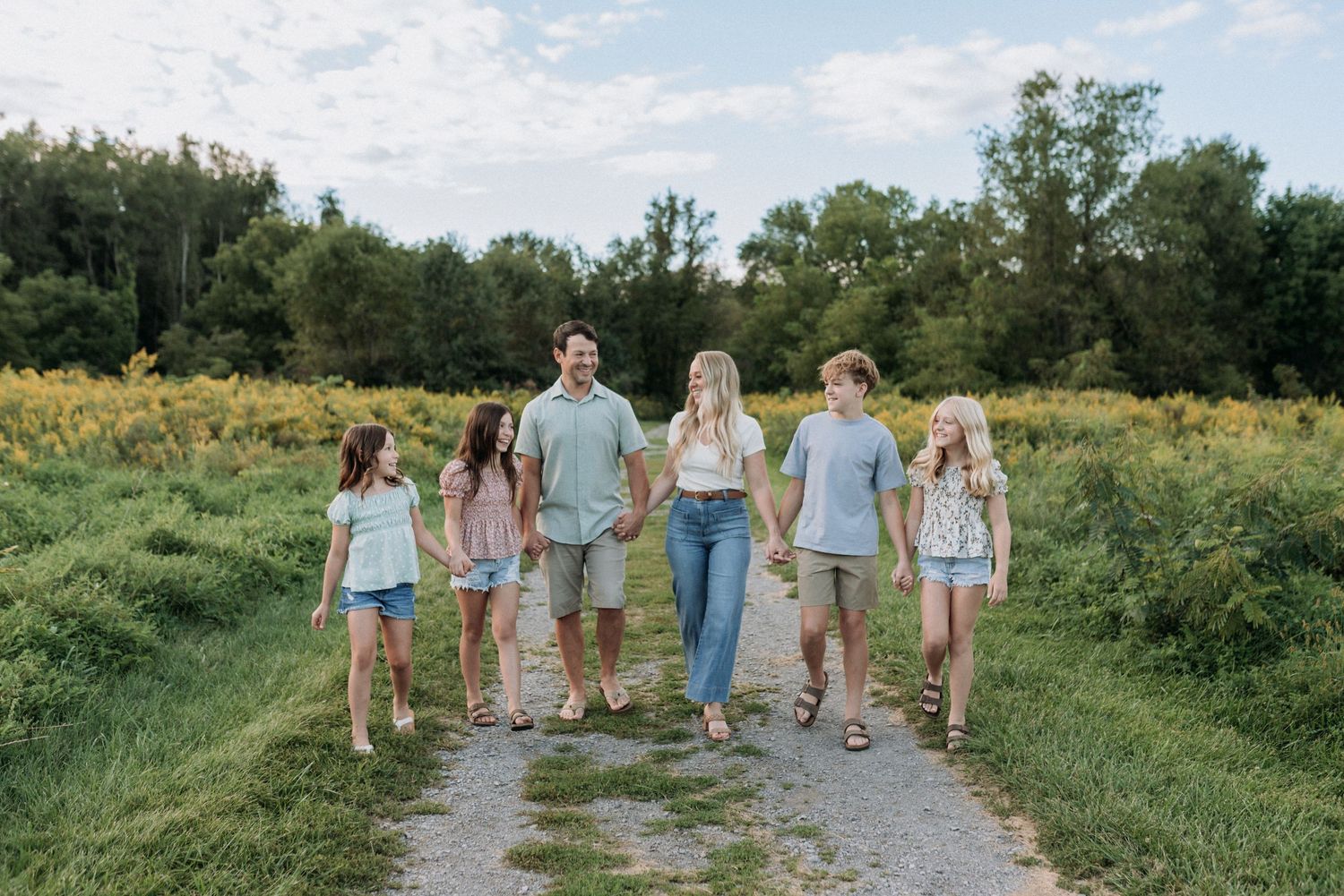 A family of six walks hand-in-hand down a scenic path surrounded by lush green fields and trees.