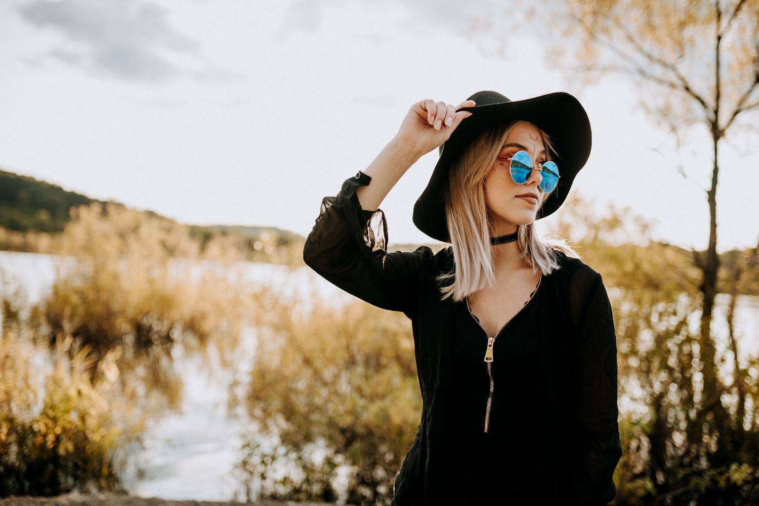 Stylish blonde woman in black outfit wearing a wide-brim hat and blue mirrored sunglasses outdoors. Senior Portrait Photography.