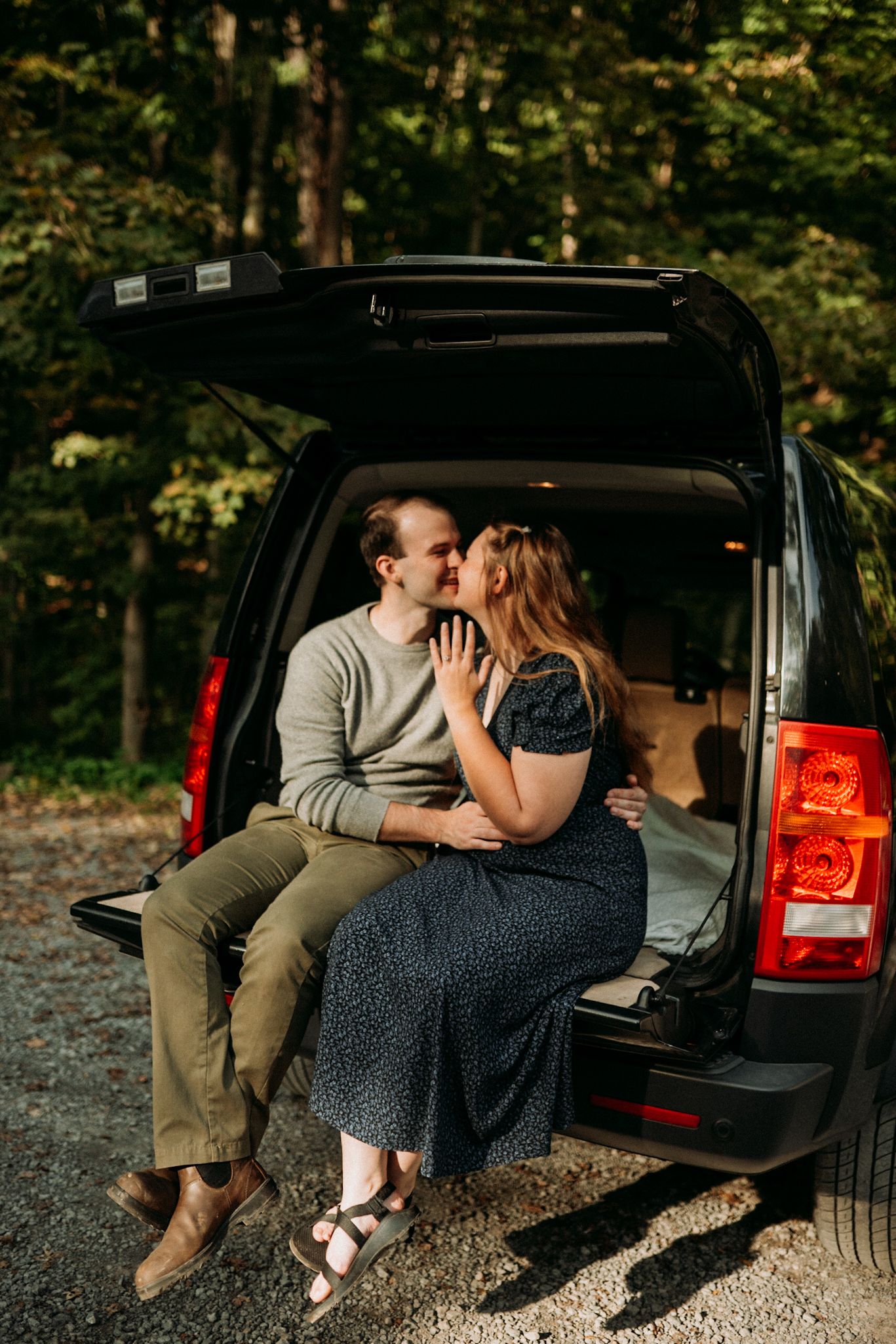 Couple sharing a kiss while sitting in open trunk of black SUV in forest. Engagement Session.