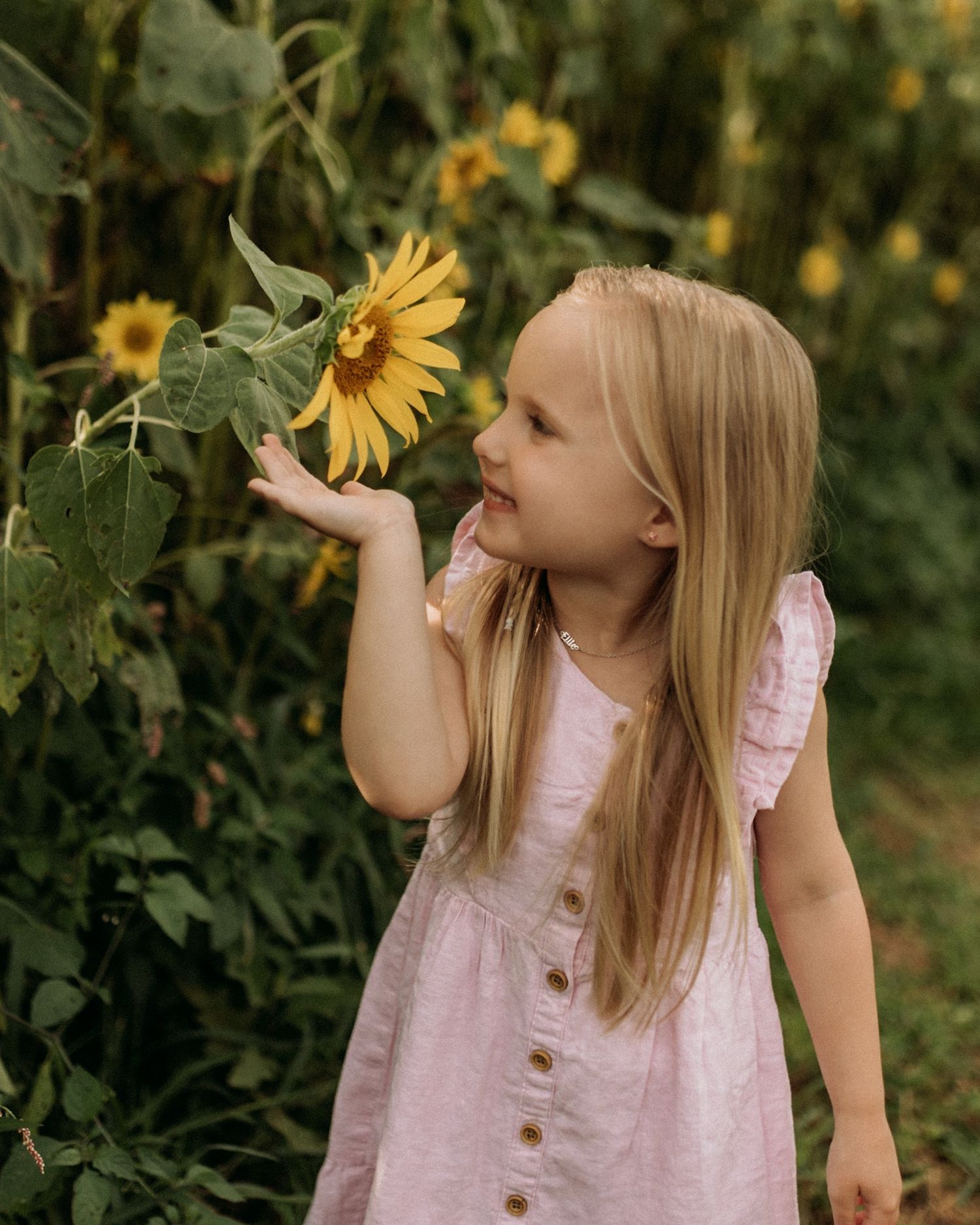 A young blonde girl in a pink dress gently touches a bright yellow sunflower in a lush garden.